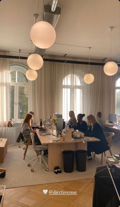 a group of people sitting around a wooden table in an open room with lots of windows