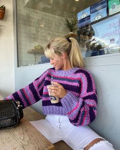 a woman sitting on a bench holding a cup and looking at her cell phone while wearing a purple striped sweater