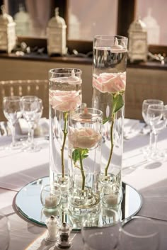 three roses in vases on a table with wine glasses and other glassware around it