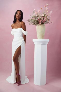 a woman in a white dress standing next to a vase with flowers on the side