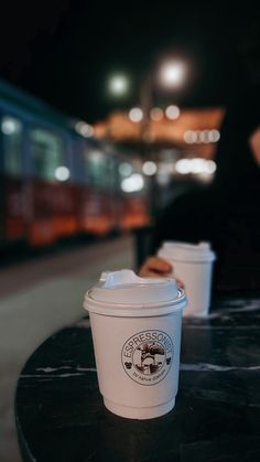 two coffee cups sitting on top of a black table next to a subway train at night