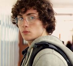 a young man with curly hair and glasses looks at the camera while wearing a backpack