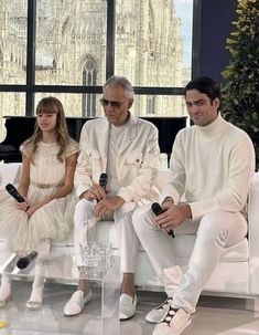 three people sitting on a white couch in front of a window with a christmas tree