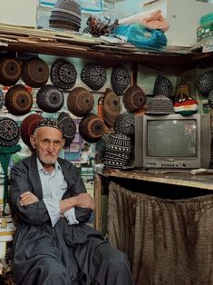 an old man sitting in front of a tv surrounded by baskets on the wall and ceiling