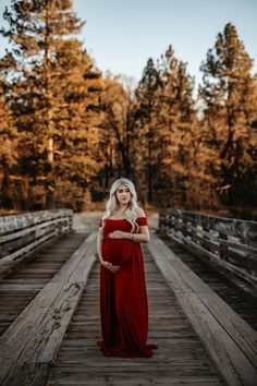 a pregnant woman in a red dress standing on a wooden bridge with trees behind her