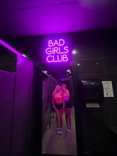 two women standing in front of a purple neon sign that says bad girls club on it