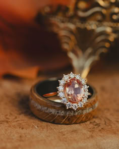 A matching wedding ring set from Staghead Designs sits on a book cover with autumn leaves and a gold jewelry case in the background. On the bottom is a black zirconium "Remmy" men's wedding band with whiskey barrel oak overlay and an offset antler inlay. Above sits the "Malina" engagement ring in 14K yellow gold with an oval cut Oregon sunstone center stone and diamond halo. Sunstone Ring Engagement, Orange Moonstone Gemstone Ring, Sunstone Rings, Oregon Sunstone Engagement Ring, Sunstone Engagement Ring, Elegant Sunstone Jewelry, Round Shape, Elk Antler