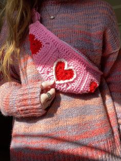 a woman wearing a pink and red sweater holding a knitted heart shaped purse in her hands