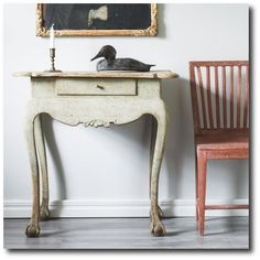 an old table with a duck on top next to a wooden chair and a painting