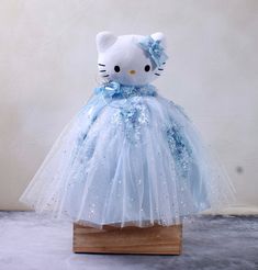 a hello kitty doll in a blue dress on a wooden stand with white walls behind it