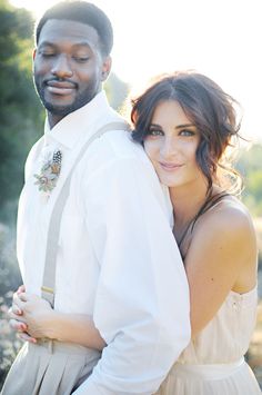 a man and woman standing next to each other in front of some trees with their arms around each other