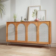 a wooden cabinet with two pictures on top and plants in the corner next to it