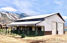 a white barn sitting on top of a dirt field next to a green fence and snow covered mountains