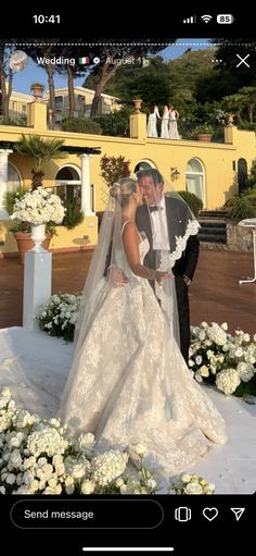 a bride and groom kissing in front of a yellow building with white flowers on the ground