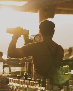 a man standing in front of a bar pouring something into a glass with the sun behind him