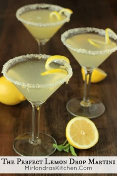 three martinis with lemon garnish sit on a wooden table next to sliced lemons