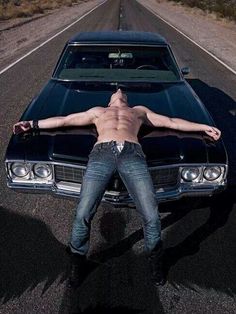 a shirtless man laying on the hood of a black car in the middle of an empty road