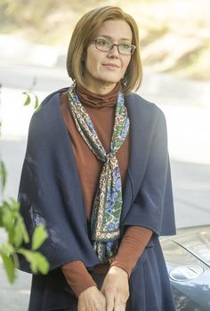 a woman wearing glasses and a scarf standing in front of a car with her hands on her hips