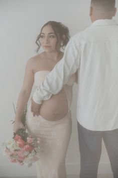 a pregnant woman in a white dress holding a bouquet and standing next to a man