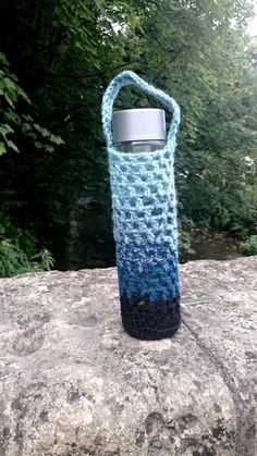 a blue and black crocheted water bottle cover sitting on top of a rock