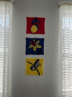 four different colored pieces of cloth hanging on the wall in front of two windows with sheer curtains