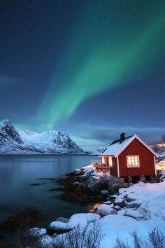 a red house sitting on top of a snow covered hillside next to the ocean with an aurora bore in the sky