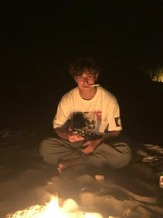 a young man sitting in front of a campfire at night with a book on his lap