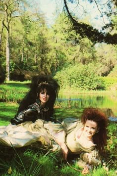 two women laying on the ground in front of a body of water with trees behind them