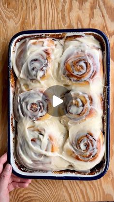 a pan filled with cinnamon rolls on top of a wooden table next to a person's hand