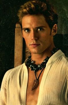 a young man wearing a white shirt and necklace with a wooden chair in the background