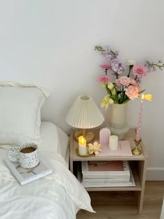 a nightstand with flowers and candles on it next to a white bed in a bedroom