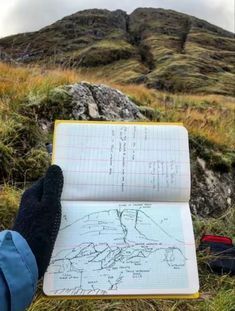 someone is writing on a notebook in the grass with mountains in the backgroud