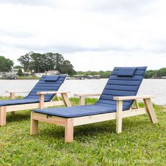 two blue chaise lounge chairs sitting on the grass near water and trees in the background