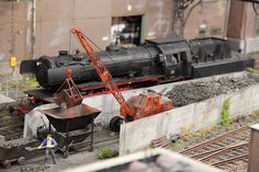 there is a model train on the tracks and people are standing next to it in front of an old brick building