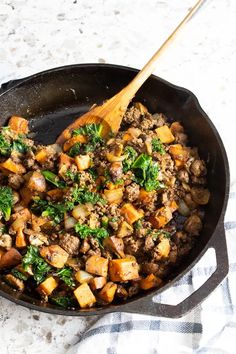 a skillet filled with meat and vegetables on top of a white table cloth next to a wooden spoon