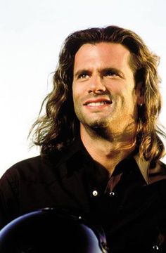 a man with long hair wearing a black shirt and holding a motorcycle helmet on his shoulder