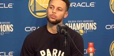 a man sitting at a microphone in front of a blue wall with the golden state warriors on it