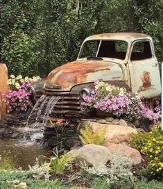 an old truck is sitting in the middle of some flowers and rocks with water coming out of it