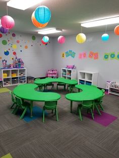 an empty classroom with green tables and colorful balls hanging from the ceiling in front of children's bookshelves