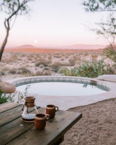 two coffee cups sitting on top of a wooden table next to a pool in the desert