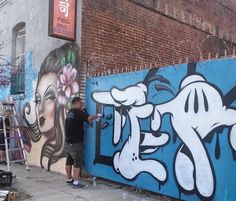 a man standing in front of a wall with graffiti on it and another person painting the side