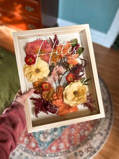 a person holding up a card with flowers on it in front of a bed and dresser