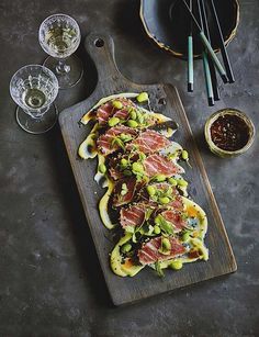 a wooden cutting board topped with food next to bowls and chopsticks on top of a table