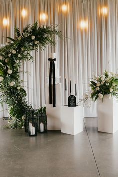 two white vases with flowers and greenery are on display in front of a curtain