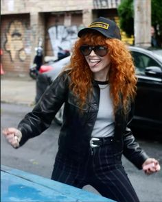 a woman with long red hair wearing sunglasses and a leather jacket is walking down the street