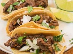 three tacos with meat, onions and cilantro on a white plate next to lime wedges