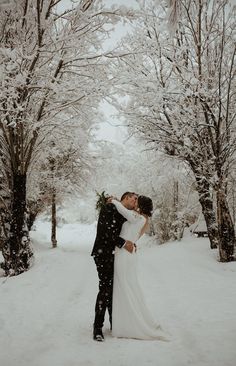 a bride and groom kissing in the snow surrounded by trees with white flowers on their wedding day