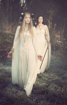 two women dressed in white are walking through the woods with long hair and holding wands