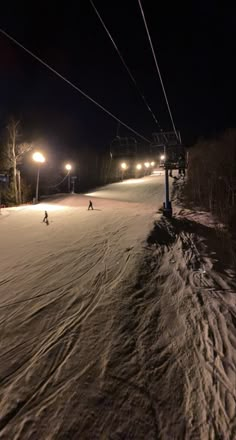 two people skiing down a snow covered slope at night with ski lift in the background