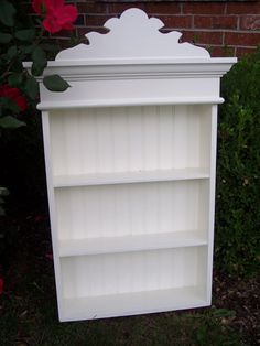 a white bookcase sitting in the grass next to a brick wall and red flowers